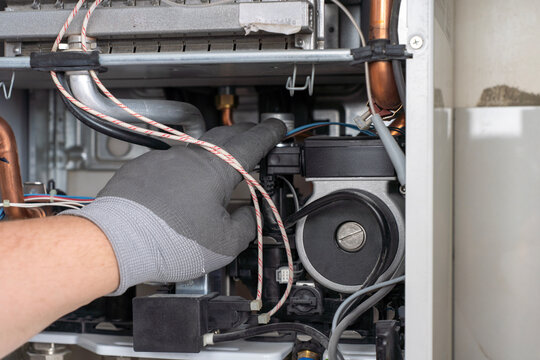 Close-up of a technician hand in a protective glove inspecting a circulation pump and hydraulic components inside an open domestic gas boiler during maintenance.