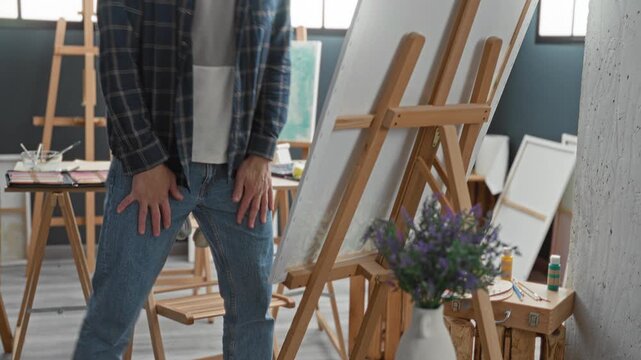 Man sitting at easel, hands clasped on knees, looking intently at canvas amid paint tools, palettes and stacked canvases in studio; creative contemplation.