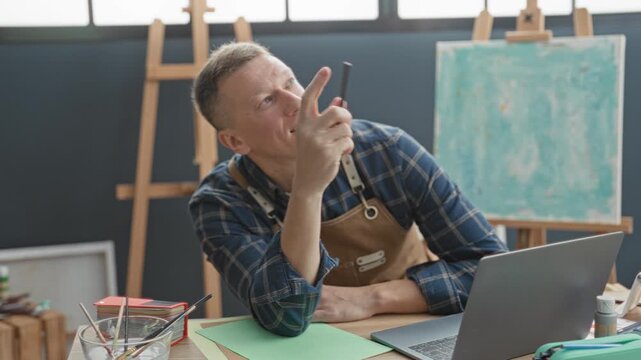 Man painter in apron holds paintbrush to chin and gazes upward in studio with laptop and easel nearby; thoughtful focus.