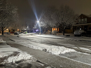Obraz premium A quiet suburban neighborhood street on a cold winter night. Bright streetlights illuminate the snow-covered road, sidewalks, and bare trees. Parked cars and houses sit silently under the dark sky.