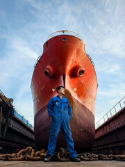 A shipyard worker stands proudly in front of a giant cargo ship under maintenance in a dry dock. © yortzafoto