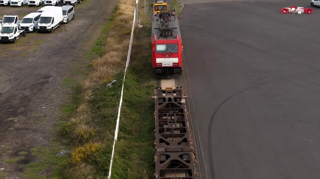 Aerial drone follows red locomotive aproaching empty flatcars past rows of white vans at industrial freight yard.