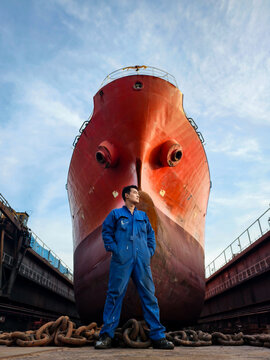 A shipyard worker stands proudly in front of a giant cargo ship under maintenance in a dry dock.
