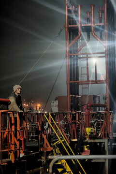 Worker stands on the deck of an offshore oil rig at night, gazing up at the lit-up derrick structure.