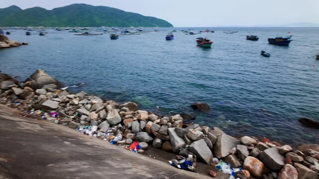 Groups of boats are seen on the water in Dak Lak, Vietnam. Rocks line the shore while some trash is present on the land. The time appears to be dusk.