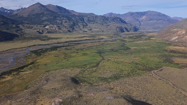A sweeping aerial view of a braided river winding through a broad Patagonian valley, framed by rugged mountain peaks, open grasslands, and rolling foothills under clear skies.