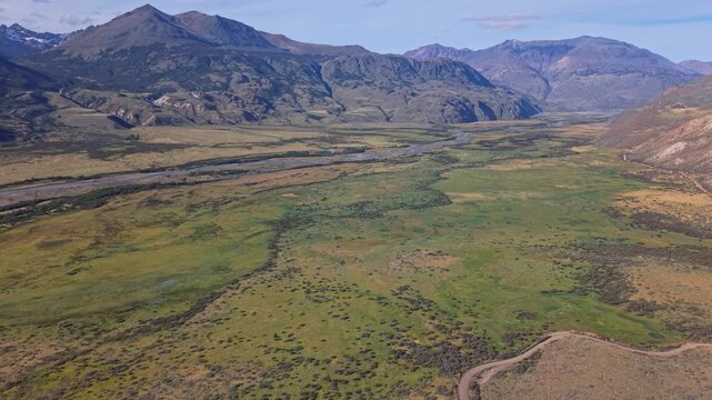 Aerial view of a braided river winding across the Patagonian valley floor, surrounded by rugged mountain ranges, open grasslands, and scattered shrubs beneath clear skies.