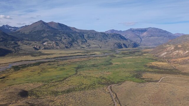 Aerial view of a braided river crossing a vast Patagonian valley, surrounded by rugged mountains, open grasslands, and untouched wilderness under clear daylight.