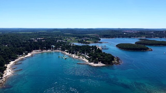 Forested islands dot sparkling deep blue Adriatic bay along Istria coastline in Croatia, colorful inflatable water park floating near sandy beach summer camping resort. Aerial drone approaching Shot
