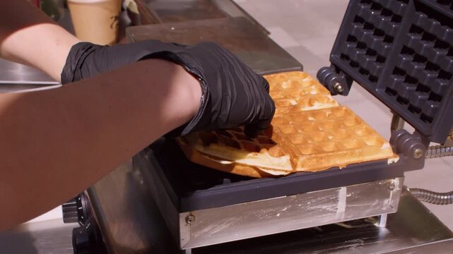 Culinary catering. Cooking occupation. Unrecognizable chef man hands in gloves opening automatic machine cutting fresh square baking waffle in restaurant.
