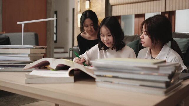 Two Asian teenage tutorials, girl concentrates on learning class at home with pile of homework, her friend uses mobile phone on sofa, self-study with textbooks, homeschooling together lifestyle.