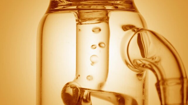 Glassy transparent liquid flowing through a glass tube, causing bubbles to condense and chaotically move inside it on yellow ramp background | Orange hydrosol cosmetic composition concept