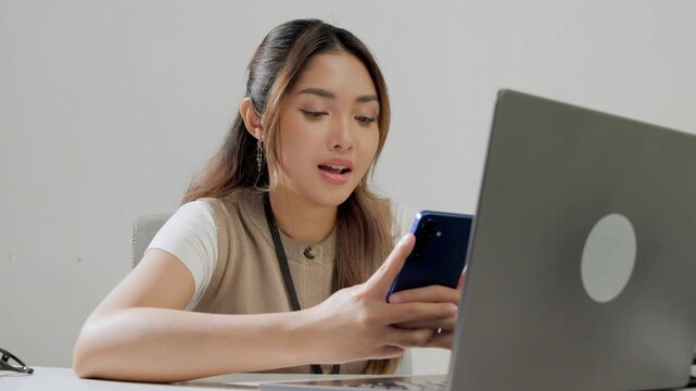 Cheerful young Asian woman is working while using smartphone to text someone or open social media.