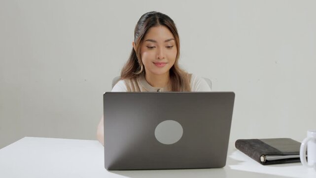 Focused young Asian woman is typing report on her laptop while feeling happy.