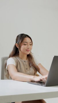 Focused young Asian woman is typing report on her laptop while feeling happy.