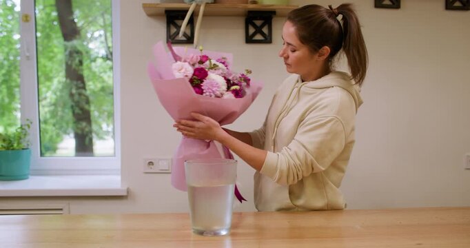 Professional floristry. Flower care. Female floral designer placing color bouquet wrapped in pink paper in glass with water in workshop indoor.
