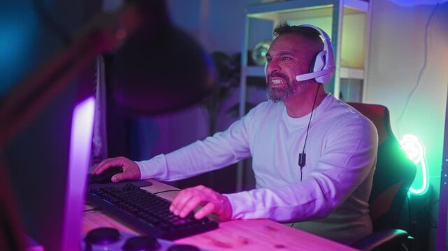 Middle-aged man wearing headset reaches with one hand toward keyboard and mouse while leaning back and closing eyes in a neon lit studio; engagement.
