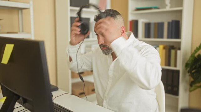 Man wearing white shirt and headset speaking into microphone at computer in building as call center agent; customer support concentration.