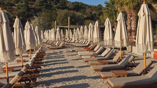 Static wide shot of rows of empty wooden sunbeds and closed boho fringe umbrellas on a sandy beach at a luxury coastal resort during peaceful morning golden hour.