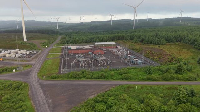 Wide View of Electrical Substation Managing and Re-Directing Flow of Electricity on Natural Landscape with Wind Turbines in Background.