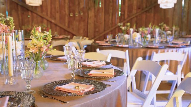 Charming rustic barn wedding reception setup with pastel floral centerpieces, glassware, metallic chargers, and peach napkins on round tables in warm, natural lighting. Stylish and inviting.