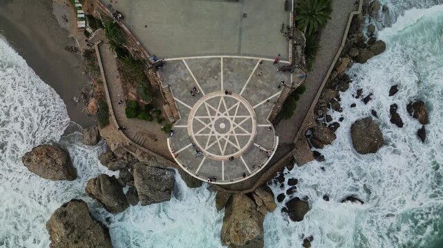Straight down drone view of Balcon de Europa circular viewpoint with star mosaic pavement surrounded by crashing waves and rocky cliffs, tourists visible, overcast daylight