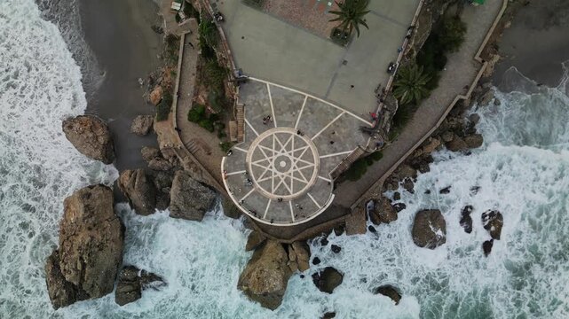 Straight down aerial drone view of Balcon de Europa circular viewpoint with star mosaic pavement, tourists walking, rocky cliffs and crashing waves surrounding the promontory, overcast daylight