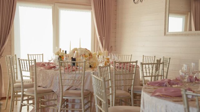 Beautifully decorated wedding reception venue with round tables, blush pink napkins, floral centerpieces, glassware, and natural light, captured in a wide angle static shot.