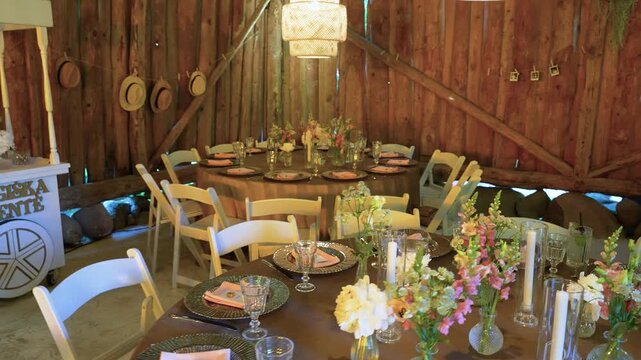 Warmly lit rustic barn venue features round tables, elegant table settings, floral centerpieces, vintage hats, glassware, and cozy string lights, perfect for a wedding or special event.