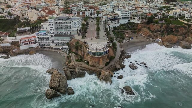 Aerial drone pullback shot with camera tilting up to reveal Nerja town center, Balcon de Europa viewpoint, palm-lined promenade and green mountains in background, overcast daylight