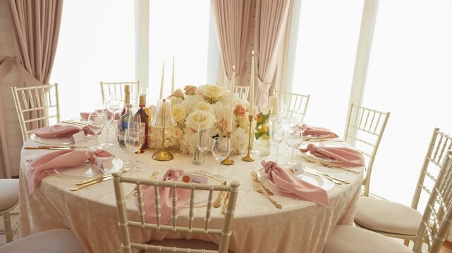 A beautifully decorated wedding reception table features blush pink napkins, gold cutlery, floral centerpiece, and candles. Soft natural light floods the room, creating a warm, inviting atmosphere.
