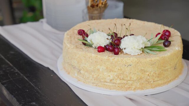 Delicate homemade cake adorned with fresh cherries and cream flowers sits on a rustic table, bathed in soft natural light. Subtle camera pan highlights texture and artisanal details.