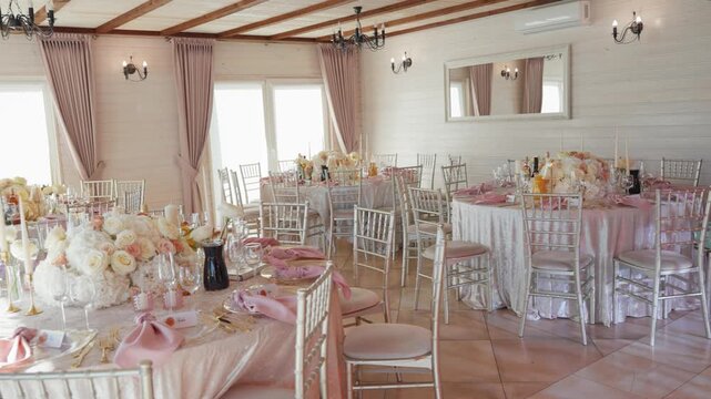 Bright banquet hall with exposed wood beams, large windows, and chandeliers, set for a formal event. Pink tablecloths, floral centerpieces, and natural light create a romantic atmosphere.