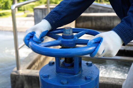 Industrial worker in protective gloves operating large blue manual valve at water treatment plant, professional maintenance engineer controlling flow in infrastructure facility, close-up, high quality