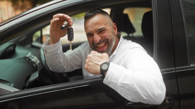 Middle age man holding car keys with raised hand and fist pump while leaning from a car window on street, smiling broadly showing teeth, white shirt and wristwatch visible; joy success.