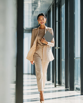 Confident successful Asian businesswoman walking through a modern glass office carrying a laptop, elegant beige business suit, warm natural sunlight, luxury corporate headquarters
