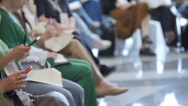Audience members sit patiently in rows during a conference. Some focus on notes or phones. Others appear relaxed or engaged. Suitable for business events or educational settings