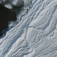 Obraz premium Aerial view of white snow ripples on frozen lake ice surface next to dark open water in a winter landscape.