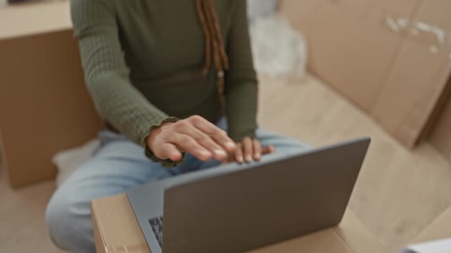 Woman typing on a laptop while sitting on stacked moving boxes inside a building during unpacking, hands on keyboard and looking down; settling calm.