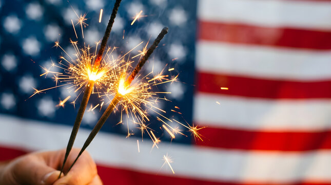 A hand holding burning sparklers against an American flag background. This image symbolizes patriotic celebration and Fourth of July festivities.