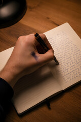 Left handed writer using fountain pen to write thoughts in paper journal at night desk.