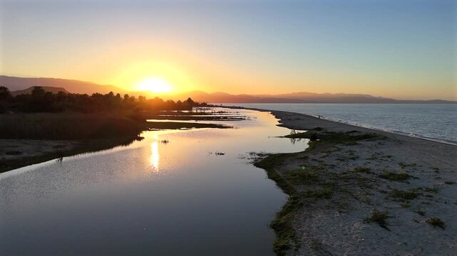 Low drone flight across the calm waters of the La Ribera estuary, approaching the natural sandbar as vibrant golden hour colors reflect across the water beneath a glowing sunset and layered desert mou