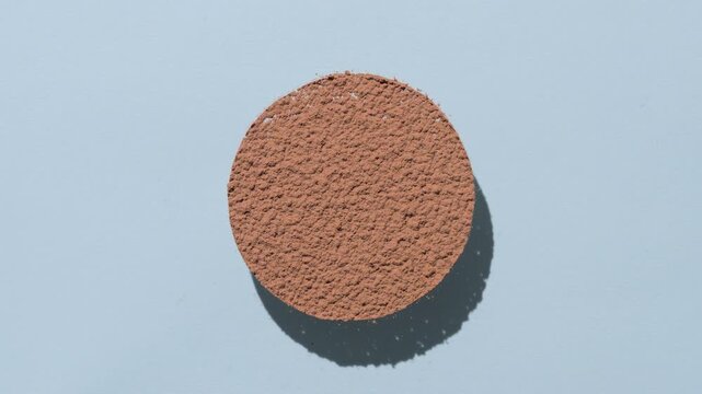 Top view perspective shot of brown powder sifted into a circle casting a morphing shadow on a faint blue background | Commercial idea for powder foundation