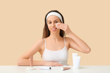 Naklejka premium Beautiful young woman with cotton pad and tubes of face cream on table against beige background
