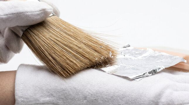 Gloved hand using wide bristle brush to apply delicate silver leaf foil