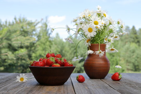 A clay bowl filled with fresh, ripe strawberries stands next to a bouquet of daisies in a clay jug, surrounded by a summer forest landscape under a blue cloudy sky.