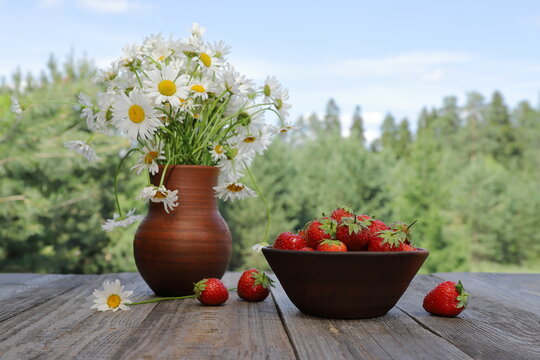 A clay bowl filled with fresh, ripe strawberries stands next to a bouquet of daisies in a clay jug, surrounded by a summer forest landscape under a blue cloudy sky.
