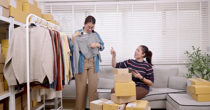 Asian women small business partners coordinate order packing activity, reviewing apparel choice, organizing online shopping parcel box workflow, managing home based e-commerce order together