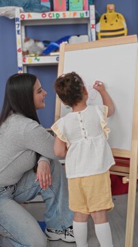 Teacher kneels beside little girl drawing on whiteboard easel with purple marker in school building; nurturing learning.