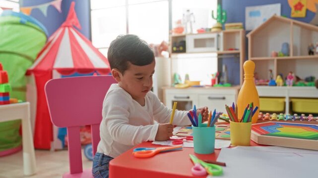 Boy holding crayon with hand drawing at a table in a colorful playroom inside a building; curiosity concentration.